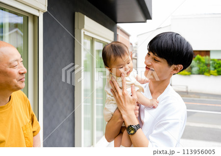 屋外の家の前で0歳児の赤ちゃんを抱っこする父親とそれを見守る祖父 113956005
