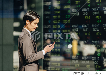 屋外の株価ボードの映る電光掲示板を前にスマートフォンでニュースを確認する30代のビジネスマン投資家 113956176