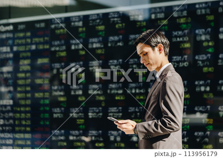 大量の株の銘柄が表示された株価ボードの前でスマートフォンを操作する30代の日本人の男性 113956179