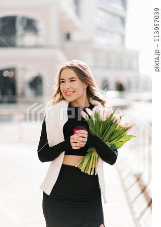 Stylish young woman with a bouquet of tulips outdoors and holding a paper cup with coffee in her hands. Portrait of a spring street lifestyle 113957039