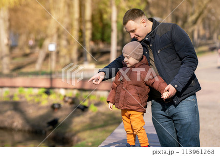 Father and toddler son stand in a park near the city lake on a sunny spring day. Family time lifestyle concept. Spending time 113961268