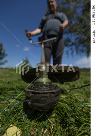 Vertical. On a clear day, a man mows the grass in his backyard with a trimmer. Hold the trimmer firmly. a man mows tall, thick grass in his backyard. A man tidies up the lawn behind his house. 113962266