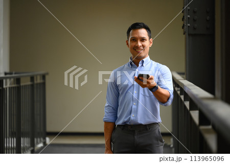 Confident businessman using mobile phone standing in the office corridor Confident businessman using mobile phone standing in the office corridor 113965096