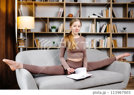 Young beautiful blonde woman in brown leggings and top sitting on a twine at home on the couch. The girl makes notes in a notebook while sitting on the sofa in an unusual position Young beautiful blonde woman in brown leggings and top sitting on a twine at home on the couch. The girl makes notes in a notebook while sitting on the sofa in an unusual position 113967803