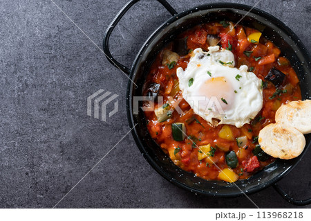 Vegetable pisto manchego with tomatoes, zucchini, peppers, onions,eggplant and egg, served in frying pan on black slate background Vegetable pisto manchego with tomatoes, zucchini, peppers, onions,eggplant and egg, served in frying pan on black slate background 113968218
