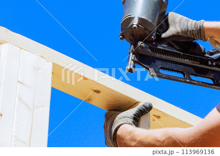 Worker in holding plank installing it on household construction using air hammer in nailing wooden beams. 113969136