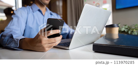 Young Asian business man executive holding cellphone device using mobile cell phone looking at smartphone working in corporate office with laptop 113970285