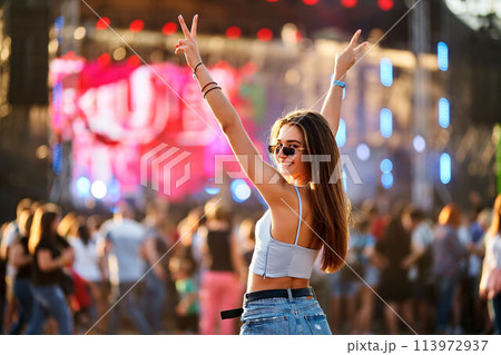 Happy young woman enjoys summer music festival on beach, arms raised, peace sign. Crowd dances near stage in sunlight. Party vibe, live concert enjoyment. Trendy outfit, sunglasses fashion statement. Happy young woman enjoys summer music festival on beach, arms raised, peace sign. Crowd dances near stage in sunlight. Party vibe, live concert enjoyment. Trendy outfit, sunglasses fashion statement. 113972937