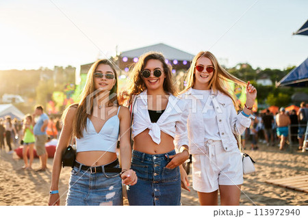 Three smiling young women walk hand in hand, sunny beach music festival. Friends in summer outfits with sunglasses, dance, socialize in coastal event. Youth culture, music on sandy shore. Three smiling young women walk hand in hand, sunny beach music festival. Friends in summer outfits with sunglasses, dance, socialize in coastal event. Youth culture, music on sandy shore. 113972940