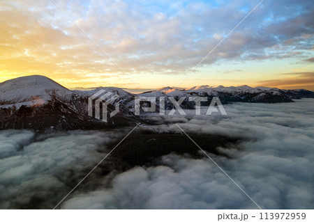 Aerial view of vibrant sunrise over white dense clouds with distant dark mountains on horizon. 113972959