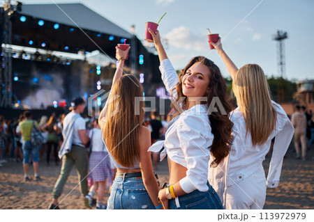 Girls with drinks enjoy live music at sunny beach festival. Group of friends dance, celebrate with cocktails. Summer party vibe on sandy shore. Attendees immerse in concert atmosphere. Girls with drinks enjoy live music at sunny beach festival. Group of friends dance, celebrate with cocktails. Summer party vibe on sandy shore. Attendees immerse in concert atmosphere. 113972979