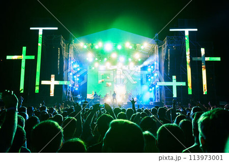 Crowd celebrates at night religious music festival with glowing stage, vibrant green lights, neon crosses. Joyful audience raises hands, enjoys live gospel performance, outdoor worship concert. Crowd celebrates at night religious music festival with glowing stage, vibrant green lights, neon crosses. Joyful audience raises hands, enjoys live gospel performance, outdoor worship concert. 113973001
