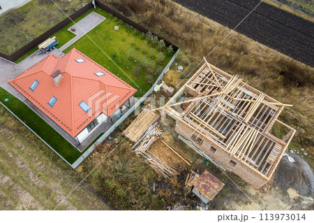 Top down aerial view of two private houses, one under construction with wooden roofing frame and another finished with red tiled roof. Top down aerial view of two private houses, one under construction with wooden roofing frame and another finished with red tiled roof. 113973014