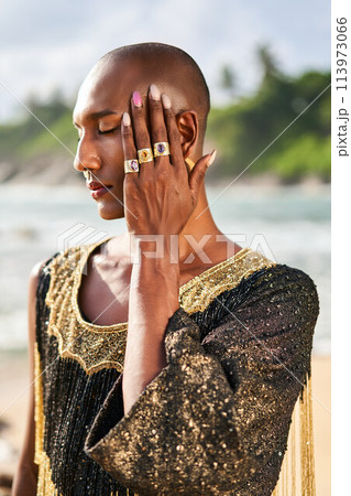 Outrageous gay black man in luxury gown, jewelry poses on scenic ocean beach. Gender fluid ethnic fashion model in posh dress, accessories looks at camera covers face with hand and rings on fingers. Outrageous gay black man in luxury gown, jewelry poses on scenic ocean beach. Gender fluid ethnic fashion model in posh dress, accessories looks at camera covers face with hand and rings on fingers. 113973066