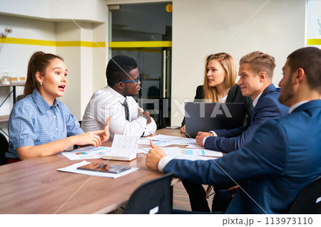 Multiracial team discusses strategy in modern office, African executive listens as Caucasian woman explains point, male CEO looks on, laptop displays charts, colleagues collaborate on project 113973110
