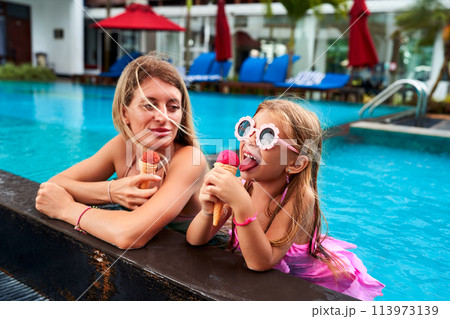 Mother, daughter enjoy ice cream by resort pool. Little girl in swim goggles eats dessert with mom on sunny day. Happy family moments, luxury vacation lifestyle. Child, parent share treat outdoors. Mother, daughter enjoy ice cream by resort pool. Little girl in swim goggles eats dessert with mom on sunny day. Happy family moments, luxury vacation lifestyle. Child, parent share treat outdoors. 113973139