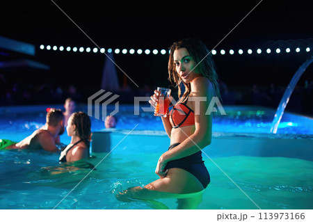 Pretty woman in bikini drinking cocktail and hanging out with friends at night pool party. People having fun dancing with drinks and splashing water in luxury private villa swimming pool. 113973166
