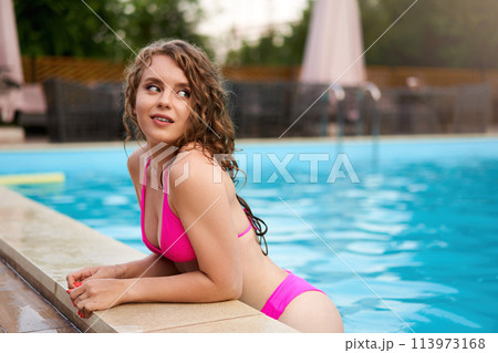 Pretty woman in pink bikini leaning at deck on pool party in beach club. Curly cute girl in swimwear chills smiling on summer holiday. Lady relaxes swimming in water in hotel resort swimming pool. Pretty woman in pink bikini leaning at deck on pool party in beach club. Curly cute girl in swimwear chills smiling on summer holiday. Lady relaxes swimming in water in hotel resort swimming pool. 113973168