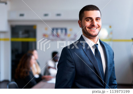 Confident eastern businessman smiles in modern office, diverse team works behind. Arab professional in formal suit leads multicultural group. Inclusive work environment, leadership, and cooperation. 113973199