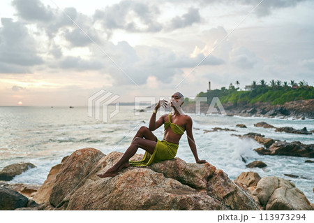 Gender fluid black person poses gracefully sitting on rocks in ocean at sunset. Androgynous slim ethnic fashion model in tropical maxi dress on top of rocky hill above storm at dusk. Pride LGBTQIA 113973294