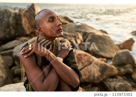 Queer black person in luxury dress, jewelry sits on rocks in ocean. Lgbtq ethnic fashion model wearing jewellery dressed in posh gown poses gracefully in tropical seaside location. Pride LGBTQIA 113973301