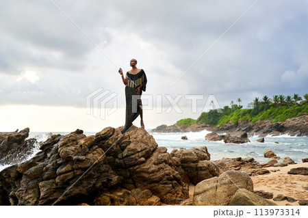 Non-binary black godlike person poses gracefully standing on rocks in ocean. Trans ethnic fashion model in posh dress and jewellery on rocky beach by storm . Lgbtq. Pride month Non-binary black godlike person poses gracefully standing on rocks in ocean. Trans ethnic fashion model in posh dress and jewellery on rocky beach by storm . Lgbtq. Pride month 113973314