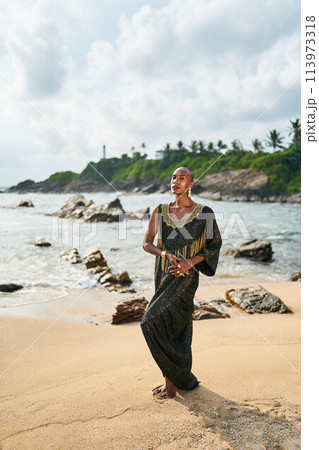 Outrageous gay black person in luxury gown poses on scenic ocean beach. Non-binary ethnic fashion model in posh outfit with makeup stands gracefully at picturesque location with sea, lighthouse. Outrageous gay black person in luxury gown poses on scenic ocean beach. Non-binary ethnic fashion model in posh outfit with makeup stands gracefully at picturesque location with sea, lighthouse. 113973318