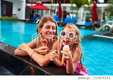 Cheerful young girl with sunglasses enjoys ice cream beside smiling mom in a pool. Relaxing family moment during sunny holiday, bonding over sweet treats, water in background, summer vibe. Cheerful young girl with sunglasses enjoys ice cream beside smiling mom in a pool. Relaxing family moment during sunny holiday, bonding over sweet treats, water in background, summer vibe. 113973331