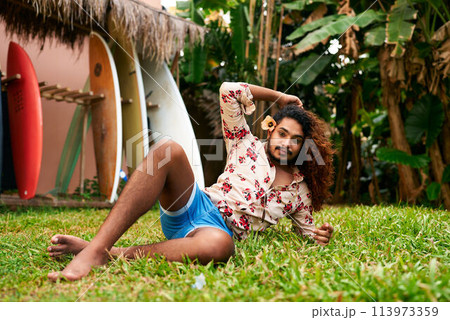 Vibrant gay man with curly hair poses in tropical garden, colorful surfboards in background. He wears floral shirt, blue shorts, outdoor fashion. LGBT traveler enjoys exotic destination, relaxed vibe. 113973359