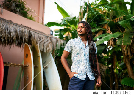 Confident dark-skinned surf instructor stands among colorful boards in tropical setting. BIPOC coach awaits students for lesson by lush green plants. Surf camp mentor ready for day training sessions. 113973360