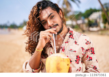 Gay man enjoys fresh coconut drink on sunny beach. Smiles, relaxed pose, tropical vacation vibe. LGBT travel lifestyle, solo male on exotic coast drinks healthy, natural beverage. Warm, clear day. 113973361