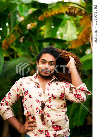 Vibrant man with rich curly hair, floral shirt stands in tropical green plants. He poses confidently, hand in hair, bold fashion, expression of LGBTQ pride and joy in nature backdrop. 113973362