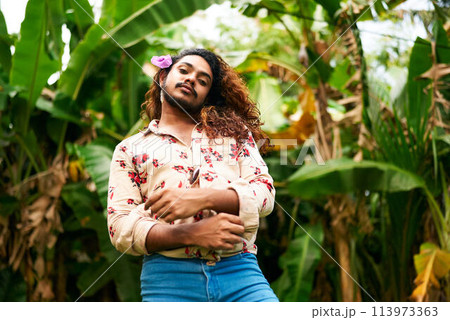 South Asian man with flower in hair stands confidently in lush garden. Expresses gay pride, embraces diversity, celebrates identity in nature. Fashionably dressed, he poses with elegance. 113973363