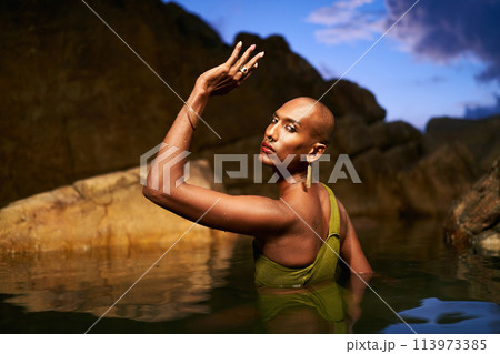 Androgynous black lgbtq fashion model dives out of water inside picturesque natural pool at night. Non-binary biethnic person poses waist deep in still water among rocks on exotic island. Pride month. Androgynous black lgbtq fashion model dives out of water inside picturesque natural pool at night. Non-binary biethnic person poses waist deep in still water among rocks on exotic island. Pride month. 113973385