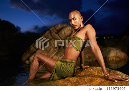 Gender fluid black person poses in creek laying on rocks in natural water pool. Queer ethnic fashion model in revealing clothes wears brass earrings, golden nose-ring, rings, bracelets lying in lake. 113973405