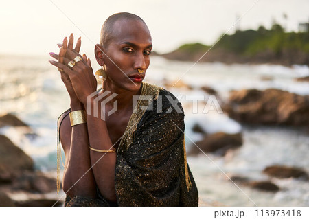 Non-binary black person in luxury dress, golden jewelry on beach rocks in ocean. Trans ethnic fashion model wearing jewellery in posh gown poses gracefully in tropical seaside location on sunset. Non-binary black person in luxury dress, golden jewelry on beach rocks in ocean. Trans ethnic fashion model wearing jewellery in posh gown poses gracefully in tropical seaside location on sunset. 113973418