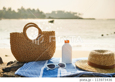 Straw bag, white sunglasses rest on blue striped towel at sunny sea shore. Summer beach day essentials hat, sunscreen bottle for skin protection. Holiday accessories by the ocean, calm water backdrop. 113973436