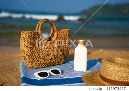 Woven bag, white sunglasses rest on blue striped towel. Bottle of sunscreen, straw hat nearby. Sandy beach setting with waves, leisure summer vibes near ocean. Travel, holiday accessories displayed. 113973437