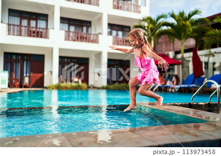 Excited young girl in pink swimsuit jumps into outdoor pool at sunny resort. Happiness, active vacation, water play depicted. Childhood, summer, leisure, travel, luxury hotel backdrop. Excited young girl in pink swimsuit jumps into outdoor pool at sunny resort. Happiness, active vacation, water play depicted. Childhood, summer, leisure, travel, luxury hotel backdrop. 113973458
