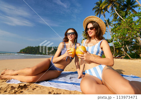 women in striped swimsuits enjoy coconut drinks on sandy beach. Sunbathers relax under tropical sun, clear blue sky. Friends on summer holiday soak up sun, sea breeze by ocean. Palm idyllic coast. women in striped swimsuits enjoy coconut drinks on sandy beach. Sunbathers relax under tropical sun, clear blue sky. Friends on summer holiday soak up sun, sea breeze by ocean. Palm idyllic coast. 113973471