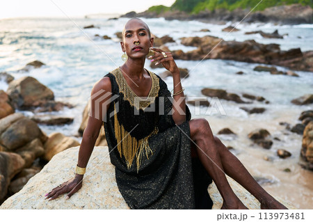 Queer black person in luxury dress, jewelry sits on rocks in ocean. Lgbtq ethnic fashion model wearing jewellery dressed in posh gown poses gracefully in tropical seaside location. Pride LGBTQIA Queer black person in luxury dress, jewelry sits on rocks in ocean. Lgbtq ethnic fashion model wearing jewellery dressed in posh gown poses gracefully in tropical seaside location. Pride LGBTQIA 113973481