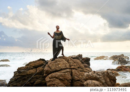 Non-binary black godlike person poses gracefully standing on rocks in ocean. Trans ethnic fashion model in posh dress and jewellery on rocky beach by storm . Lgbtq. Pride month 113973491