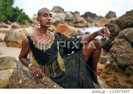 Epatage lgbtq black man poses on scenic ocean beach looks at camera demonstrates jewellery. Androgynous ethnic fashion model in posh dress, jewelry looks at camera, sits on stone. Pride month. 113973497