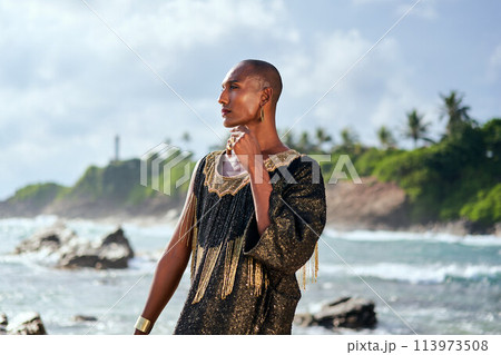 Outrageous black lgbtq person in luxury gown poses on scenic ocean beach. Non-binary ethnic fashion model in posh outfit stands gracefully at picturesque location near sea, lighthouse. Pride month. Outrageous black lgbtq person in luxury gown poses on scenic ocean beach. Non-binary ethnic fashion model in posh outfit stands gracefully at picturesque location near sea, lighthouse. Pride month. 113973508