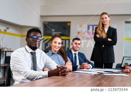 Multiethnic business team sits at meeting table in modern office. African, Caucasian, Arabic execs discuss strategy. Young woman leads diverse group. Colleagues with laptop, prepare presentation. Multiethnic business team sits at meeting table in modern office. African, Caucasian, Arabic execs discuss strategy. Young woman leads diverse group. Colleagues with laptop, prepare presentation. 113973544