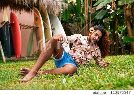 Vibrant gay man with flower in hair sits on grass, surfboards casual summer wear, relaxed. LGBT traveler enjoys tropical garden, represents diversity in leisure, confidence meet vacation bliss. 113973547