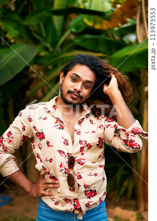 Vibrant ethnic gay man poses in botanical setting wearing floral shirt, blue jeans. Confident queer subject touches hair, stands by green plants. Representation of pride, confidence, summer fashion. Vibrant ethnic gay man poses in botanical setting wearing floral shirt, blue jeans. Confident queer subject touches hair, stands by green plants. Representation of pride, confidence, summer fashion. 113973553