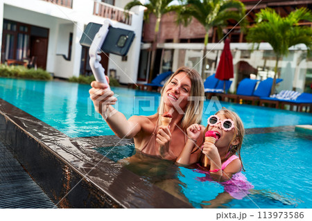 Mother and daughter enjoy ice cream while swimming in resort pool. Mom uses selfie stick for live stream video. Happy family vacation moment with tech interaction. Mom blogs, child smiles with treat. 113973586