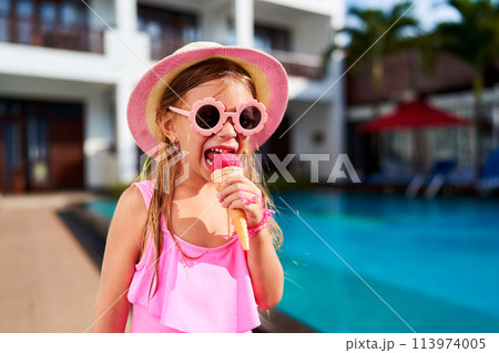 Little girl in pink swimsuit, sunhat enjoys ice cream near pool. Child cools off with sweet treat on summer day. Bright sunshine, holiday relaxation, playful kid at resort. Cheerful moment by water. Little girl in pink swimsuit, sunhat enjoys ice cream near pool. Child cools off with sweet treat on summer day. Bright sunshine, holiday relaxation, playful kid at resort. Cheerful moment by water. 113974005