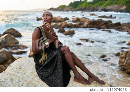 Non-binary black person in luxury dress, golden jewelry on beach rocks in ocean. Trans ethnic fashion model wearing jewellery in posh gown poses gracefully in tropical seaside location on sunset. Non-binary black person in luxury dress, golden jewelry on beach rocks in ocean. Trans ethnic fashion model wearing jewellery in posh gown poses gracefully in tropical seaside location on sunset. 113974028